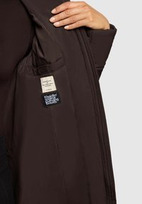 Braune isolierte Jacke mit gesteppter Innenfütterung. Mit Innenetiketten, darunter Pflegehinweise und Markenlogo. Gezeigt wird strukturierter Stoff.