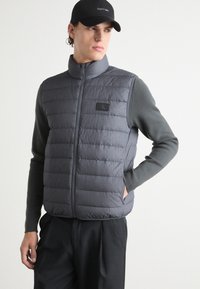 Graue gepolsterte Weste mit hohem Kragen, die vertikale Nähte und ein Markenpatch aufweist. Darunter ist ein dunkelgraues Langarmshirt sichtbar.