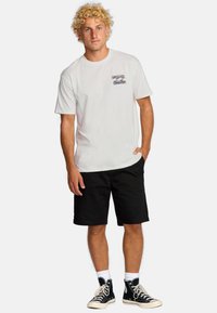 Hvid kortærmet t-shirt med grafisk print og lomme detalje, parret med sorte shorts og sorte høje sneakers.