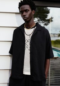 Maglietta nera a maniche corte con texture sopra una camicia bianca a pois con un disegno grafico. Il modello indossa collane a strati di perline.