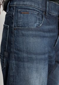 Dunkelblaue Jeans aus Denim mit einem klassischen Fünf-Taschen-Design, G-Star-Logo-Patch und subtilen Ausbleichungen auf der Stoffoberfläche.