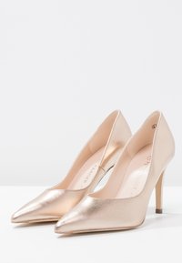 Une paire d'escarpins pointus à talons hauts de couleur or rose métallique exposée sur une surface blanche avec un fond gris clair uni.