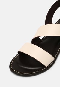 Sandale en cuir beige clair avec deux larges brides sur une semelle intérieure marron foncé. Présente une couture visible le long du bord pour un détail supplémentaire.
