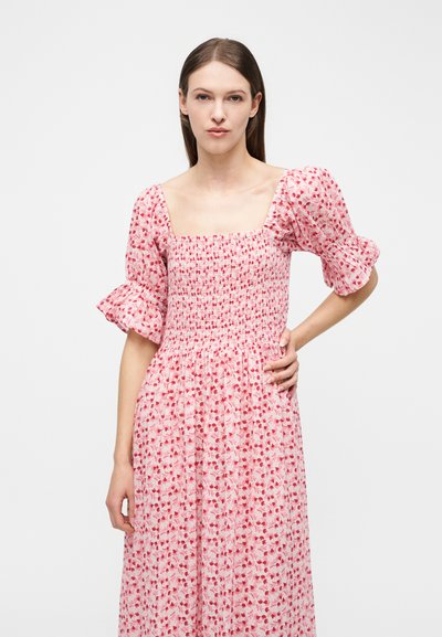 Γυναίκα με ίσια καστανά μαλλιά φοράει ροζ και κόκκινο φόρεμα με floral σχέδιο, φουσκωτά μανίκια μέχρι τον αγκώνα και τετράγωνο ντεκολτέ, με το χέρι στη μέση, σε λευκό φόντο.