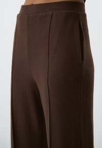 Braune weite Hosen aus strukturiertem Stoff, mit hohem Bund und scharfen Falten, ohne sichtbare Taschen oder Beschläge.