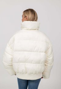 Witte pufferjack met een hoge kraag, doorgestikt ontwerp, ribgebreide zoom en oversized pasvorm, gedragen over blauwe jeans. Glad, glanzend textuur.