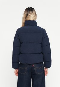 Person med langt ombre hår som bruker en marineblå dunjakke og mørk denim jeans, vendt bort fra en enkel hvit bakgrunn.