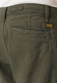 Close-up do bolso traseiro de calças de cor verde-azeitona com textura, apresentando o rótulo da marca G-Star Originals RAW Denim em preto e amarelo.