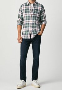 Man som bär en grön och vit rutad knappar skjorta, mörkblå jeans och vita sneakers, står med ena handen i fickan.