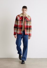 Red, plaid flannel jacket with a beige collar, worn over a light beige sweater and blue jeans. Model stands on a neutral background.