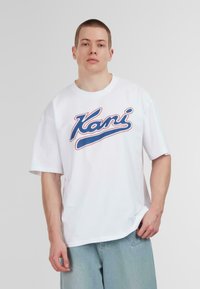 White oversized t-shirt featuring a blue and red cursive "Kani" graphic on the front. Made of cotton, it has short sleeves and a round neckline.