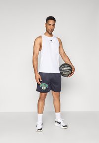Hvid atletisk tanktop, marineblå shorts med "BOSTON" grafik, holder en sort Spalding basketball, iført sort og hvidt sneakers.