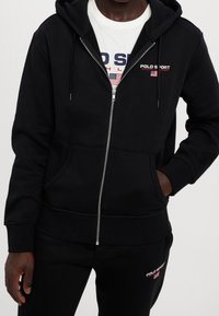 Černá mikina na zip s kapucí a šňůrkami, s předními kapsami a měkkou texturou. Je kombinována s bílým tričkem s logem "POLO SPORT".