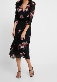 Kvinna i en svart midiklänning med omslag, dekorerad med rosa och vita blommönster samt puffärmar med trekvarts längd, ihop med svarta klackskor med remmar.