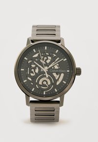 Μαύρο και ασημί μεταλλικό ρολόι χειρός Calvin Klein με καντράν σκελετού, τρεις υποκαντράν και ασημένιους δείκτες ώρας σε μεταλλικό λουράκι με κρίκους.