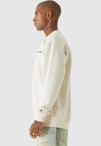 Lys beige hettegenser laget av bomull med lange ermer, ribbede mansjetter og en logo på forsiden. Satt sammen med slitte lyse denimjeans.