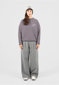 Pull gris surdimensionné avec un logo subtil sur la poitrine, associé à un large pantalon gris et des chaussures slip-on blanches. Une casquette décontractée complète le look.