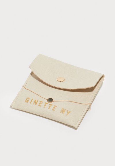 Beige műbőr tasak hajtott fedelekkel, patent záróka, arany betűzéssel "GINETTE NY", valamint egy arany lánccal és fekete medál részlettel.