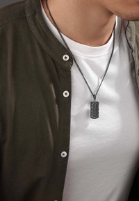 Una collana con medaglia in metallo nero pende da una catena, abbinata a una maglietta bianca e una camicia verde oliva con bottoni bianchi.