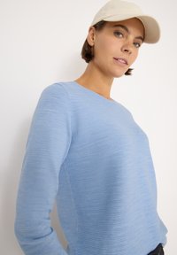 Frau, die einen hellblauen Rippstrickpullover und eine beige Mütze trägt, posiert vor einem schlichten weißen Hintergrund.