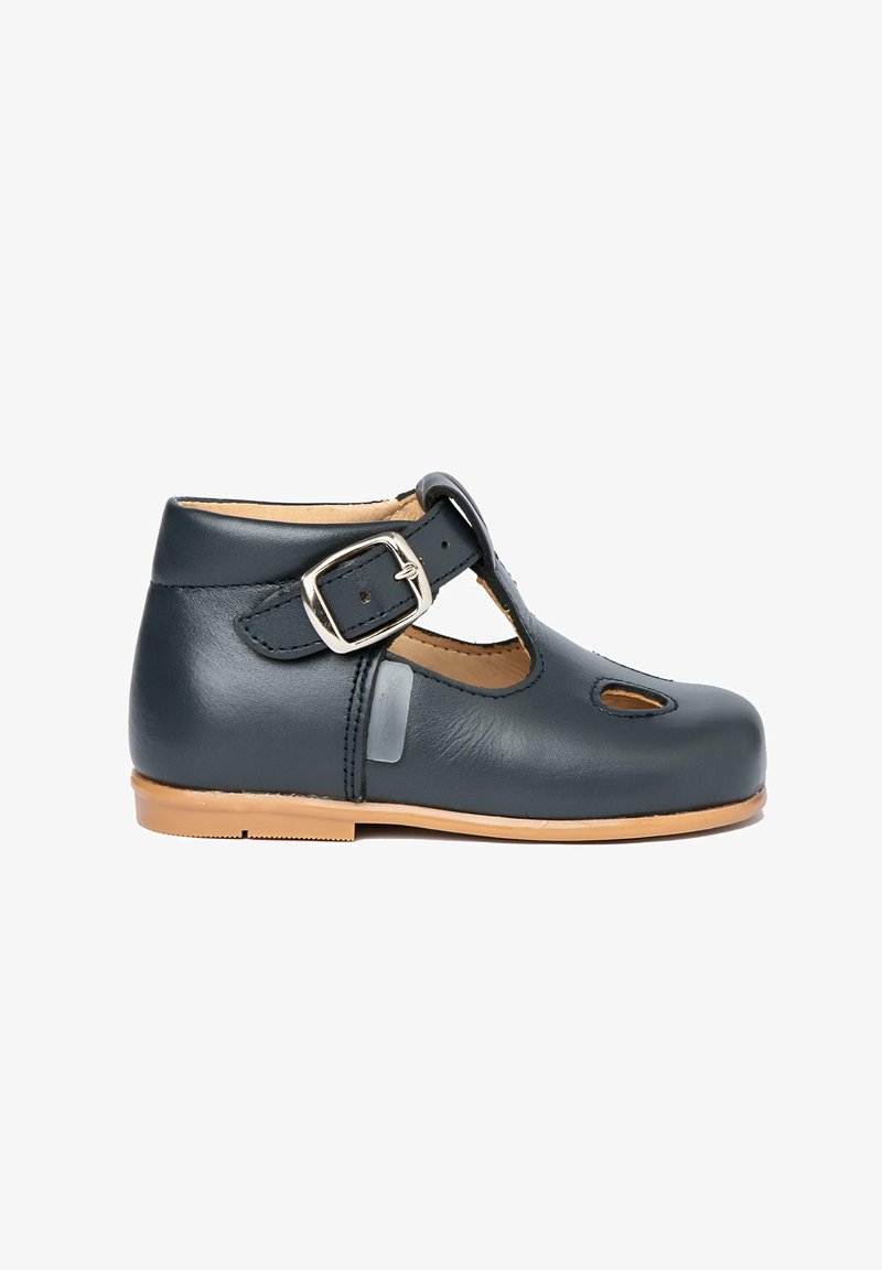 Zapato negro de cuero para niño pequeño con correa en el tobillo, hebilla plateada, pequeña apertura lateral y suela plana color beige.