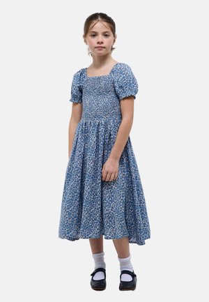 Jeune fille debout portant une robe midi bleue à fleurs avec des manches bouffantes, des chaussettes blanches et des chaussures Mary Jane noires sur un fond blanc.