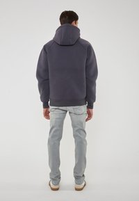 Felpa grigia scura con collo alto e cappuccio grande, dotata di polsini a coste, indossata sopra jeans azzurri chiari e sneakers beige.