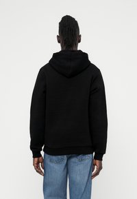 Zwarte hoodie met een gestructureerd oppervlak, rechte snit en geribbelde manchetten. Draagt een lichtblauwe spijkerbroek, gezien van achteren.