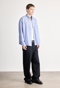 Gestreiftes blaues Hemd mit Knöpfen und Brusttasche, kombiniert mit lockeren dunklen Jeans und schwarzen Schuhen, stehend vor neutralem Hintergrund.