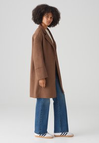 Brown wool blend oversized coat with wide sleeves, paired with blue flared jeans and white sneakers featuring navy stripes.