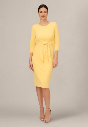 Femme debout portant une robe jaune longueur genou avec manches trois-quarts et une ceinture nouée à l'avant, associée à des talons nus à brides autour de la cheville.