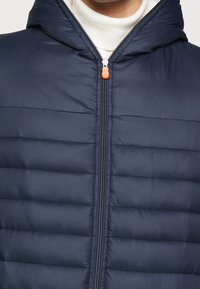 Marineblauwe pufferjack met capuchon, doorgestikt ontwerp, ritsluiting en contrasterende oranje ritstrekker. Gemaakt van een soepele, lichte stof.