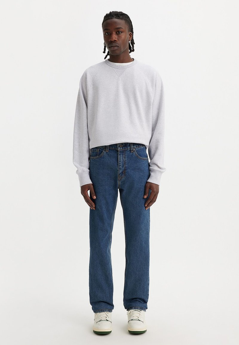 Sweatshirt gris clair à col rond et à manches longues, associé à un jean bleu à jambes droites. Des baskets blanches complètent la tenue.