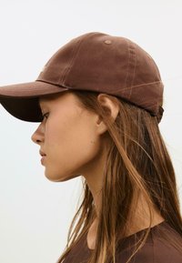 Giovane donna con lunghi capelli castani che indossa un cappellino da baseball marrone e un top marrone abbinato, voltata a sinistra su uno sfondo chiaro e uniforme.
