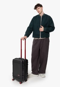 Black hard-shell suitcase with red wheels and handles, featuring a rectangular design and two zippers. Model wears a green bomber jacket and gray pants.