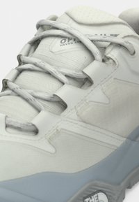 Κοντινό πλάνο ανοιχτό γκρι αθλητικού υποδήματος εξωτερικού χώρου με υφή υφάσματος, χοντρά στρογγυλά κορδόνια, επενδεδυμένη γλώσσα με κείμενο και ανθεκτική σόλα.