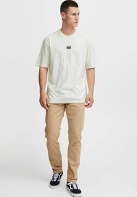 Lys gul kortærmet t-shirt med et lille sort mærke på brystet, parret med beige bukser og sorte sneakers med hvide snørrebånd.