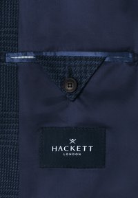 Dunkelblaues Jackenfutter mit einer zugeknöpften dreieckigen Taschenklappe und einem innen eingenähten schwarzen Hackett London Markenetikett.