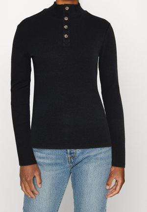 Sort ribbet langærmet top med høj krave og fire knapper, båret med lyseblå jeans på en stående person.