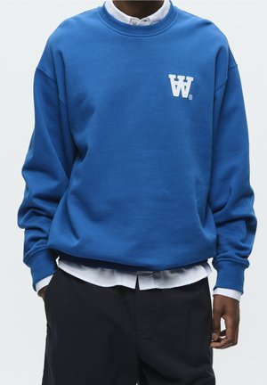 Mann trägt einen blauen Pullover mit weißem Logo auf der Brust über einem weißen Hemd und schwarzen Hosen, Hände in den Taschen.