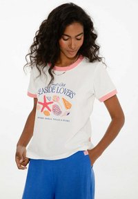 Bílý tričko s krátkým rukávem a růžovými akcenty. Na přední straně se nachází grafika "Seaside Lovers" se skořápkami a hvězdicemi; volný střih, měkký bavlněný materiál.