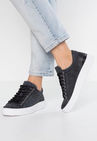 Svarta texturerade sneakers med snörningsdesign, vit sula och kontrasterande bakpanel, bärs med uppvikta ljusblå jeans.