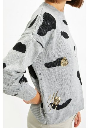 Kobieta ubrana w jasno-szary sweter z dużymi czarnymi plamami i złotymi cekinowymi akcentami, pokazana z boku z ręką na biodrze.