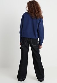 Osoba z kręconymi włosami, ubrana w granatowy, długi rękaw i czarne, wysokie spodnie typu wide-leg, widziana z tyłu.