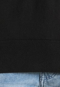 Schwarzer Strickpullover mit geripptem Saum, der ein strukturiertes Muster zeigt. Darunter befindet sich ein hellblauer Denim-Stoff, der einen lässigen, fransigen Rand hat.