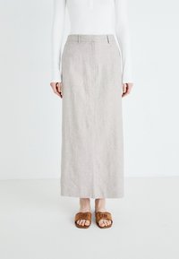 Frau trägt einen beigen langen Rock, ein weißes langärmeliges Oberteil und braune Sandalen mit offenen Zehen und steht vor einem weißen Hintergrund.