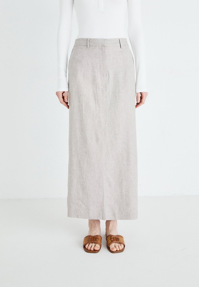 Frau trägt einen beigen langen Rock, ein weißes langärmeliges Oberteil und braune Sandalen mit offenen Zehen und steht vor einem weißen Hintergrund.