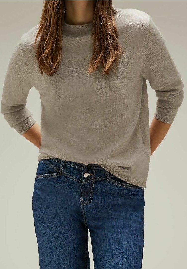Personne portant un pull beige à manches longues et un jean bleu, debout avec les mains partiellement derrière le dos devant un fond uni.