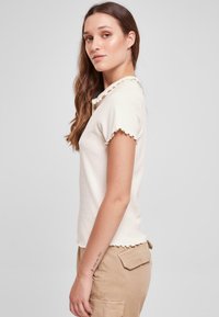 Beige kortærmet top med rynkede kanter, lavet af blød bomuld. Glat tekstur, tætsiddende design med en simpel rund halsudskæring.