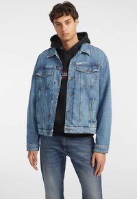 Lys blå denimjakke med klassisk snitt, to brystlommer, knappelukking og kontraststing. Brukt over en svart hettegenser.